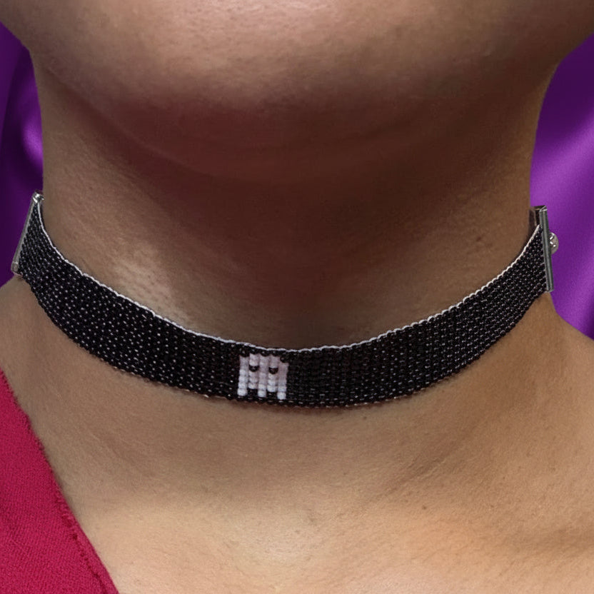 A close-up of a person wearing a black beaded choker necklace with a small silver monogram pendant. The person is wearing a red one-shoulder top in an indoor setting.