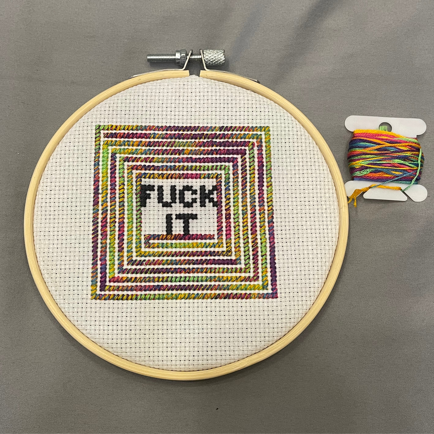 Cross-stitched hoop with 'FUCK IT' text and colorful thread on a gray background