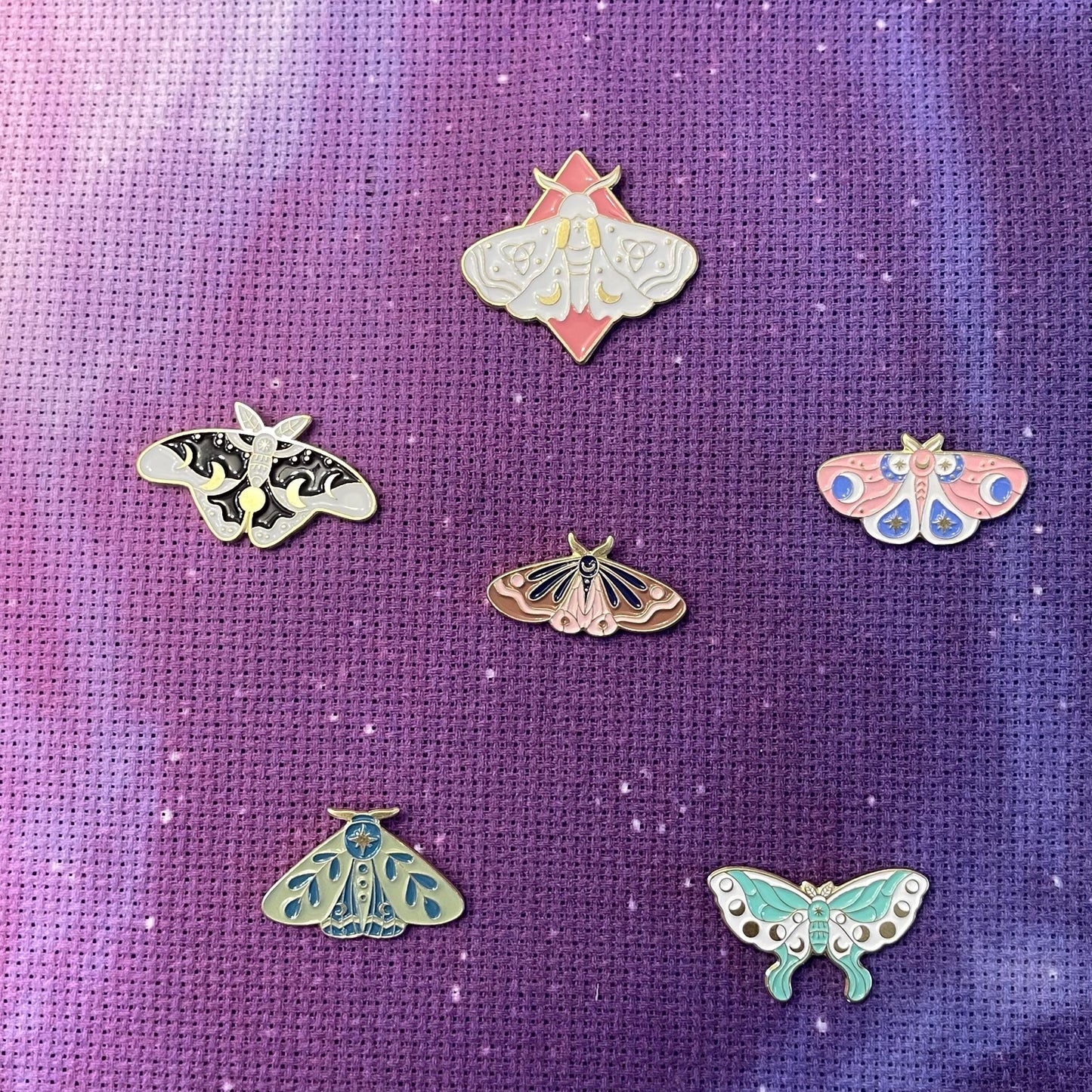 A collection of colorful enamel needle minders in the shapes of butterflies and moths displayed on a purple background.