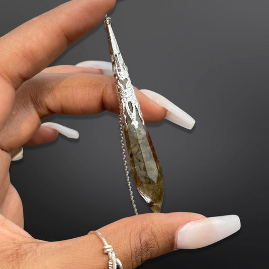 A hand holding a silver-colored labradorite pendulum with a resin encasing.