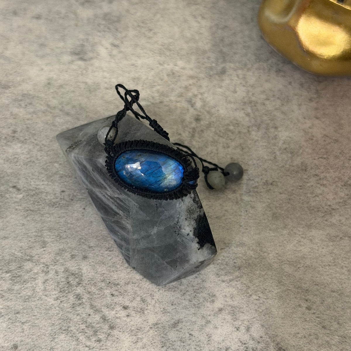 A black macrame pendant necklace with a blue iridescent moonstone cabochon, displayed on a clear quartz crystal cluster against a textured gray background.