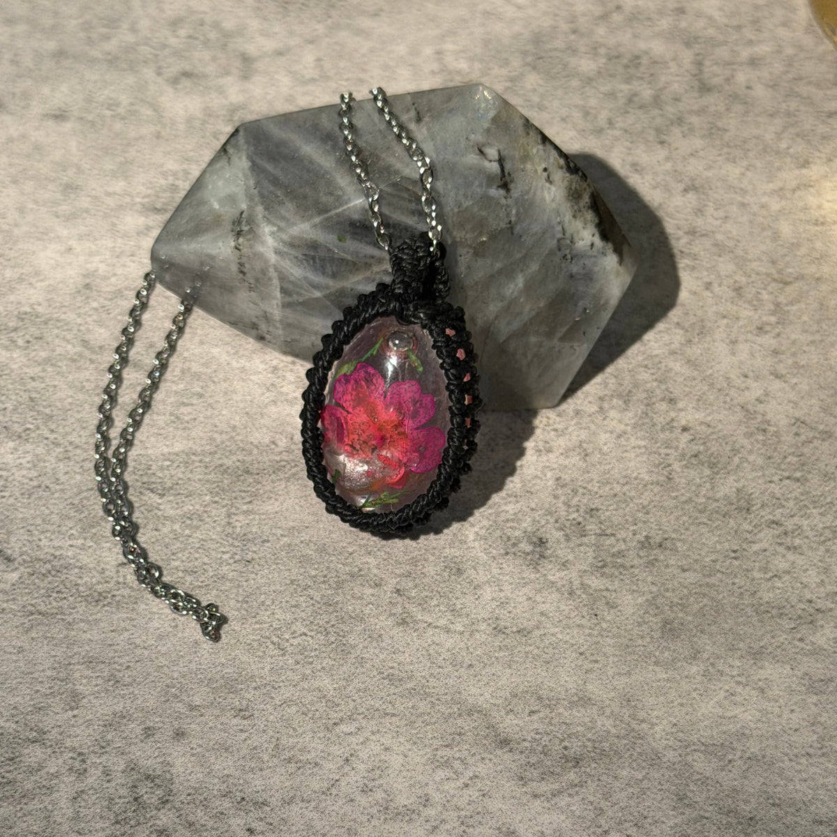 A handmade oval pendant necklace featuring a vibrant pink and red flower under clear resin, set in a black braided frame with a dark metal chain, displayed on a gray stone against a textured gray background.