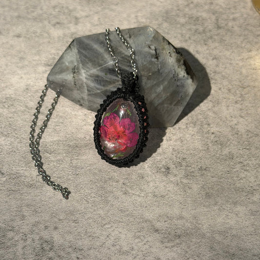 A handmade oval pendant necklace featuring a vibrant pink and red flower under clear resin, set in a black braided frame with a dark metal chain, displayed on a gray stone against a textured gray background.