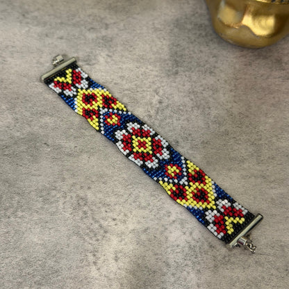 A beaded bracelet with a geometric diamond pattern in red, yellow, white, and blue, featuring silver-toned metal clasps, displayed on a textured gray surface.