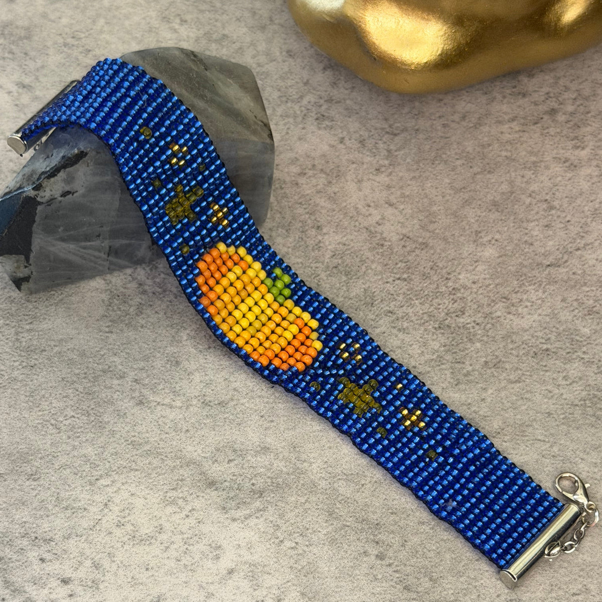 A beaded bracelet with a blue base, featuring a large orange and yellow beaded fruit motif, small yellow stars, and a silver clasp, displayed on a gray textured surface.