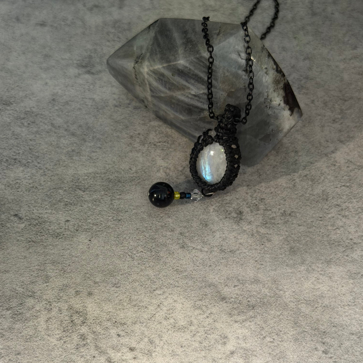 A handmade black macrame pendant necklace featuring a round iridescent moonstone and a small black bead, displayed on a textured gray surface next to a clear quartz crystal.