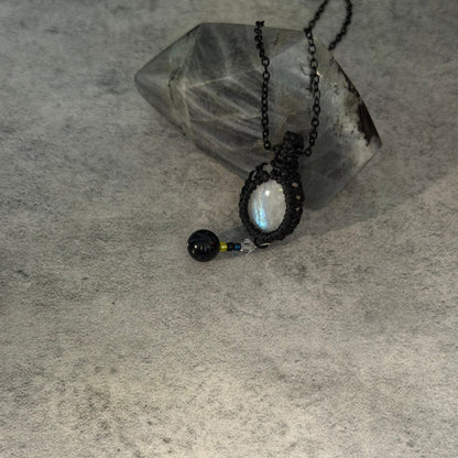 A handmade black macrame pendant necklace featuring a round iridescent moonstone and a small black bead, displayed on a textured gray surface next to a clear quartz crystal.