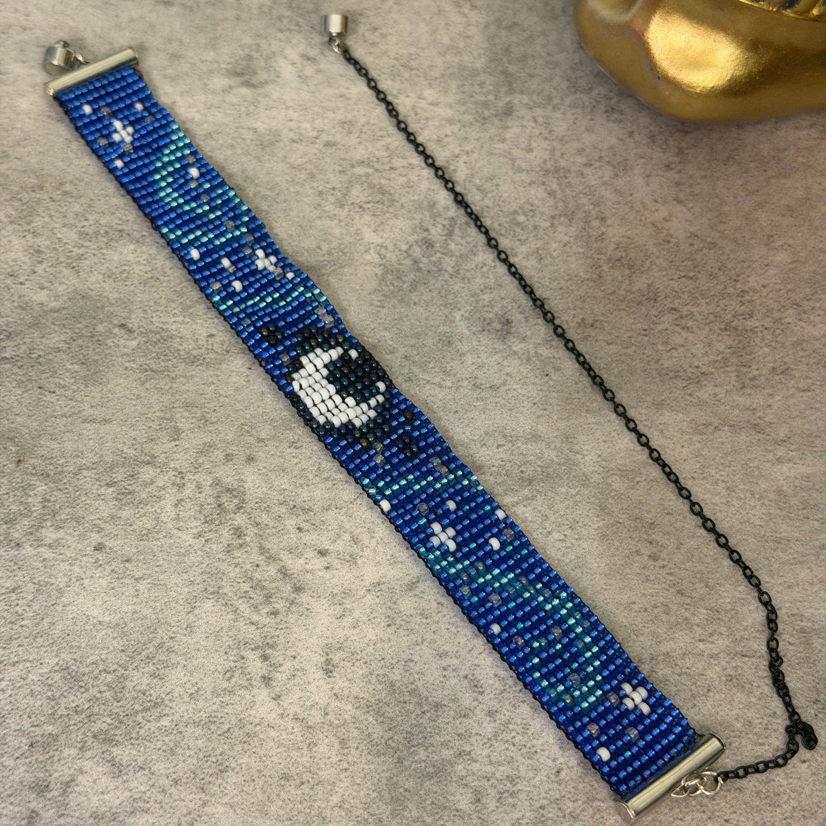 A beaded bracelet with a blue and green gradient background, white star details, and a central black-and-white crescent moon design, displayed on a gray textured surface next to a gold decorative object.