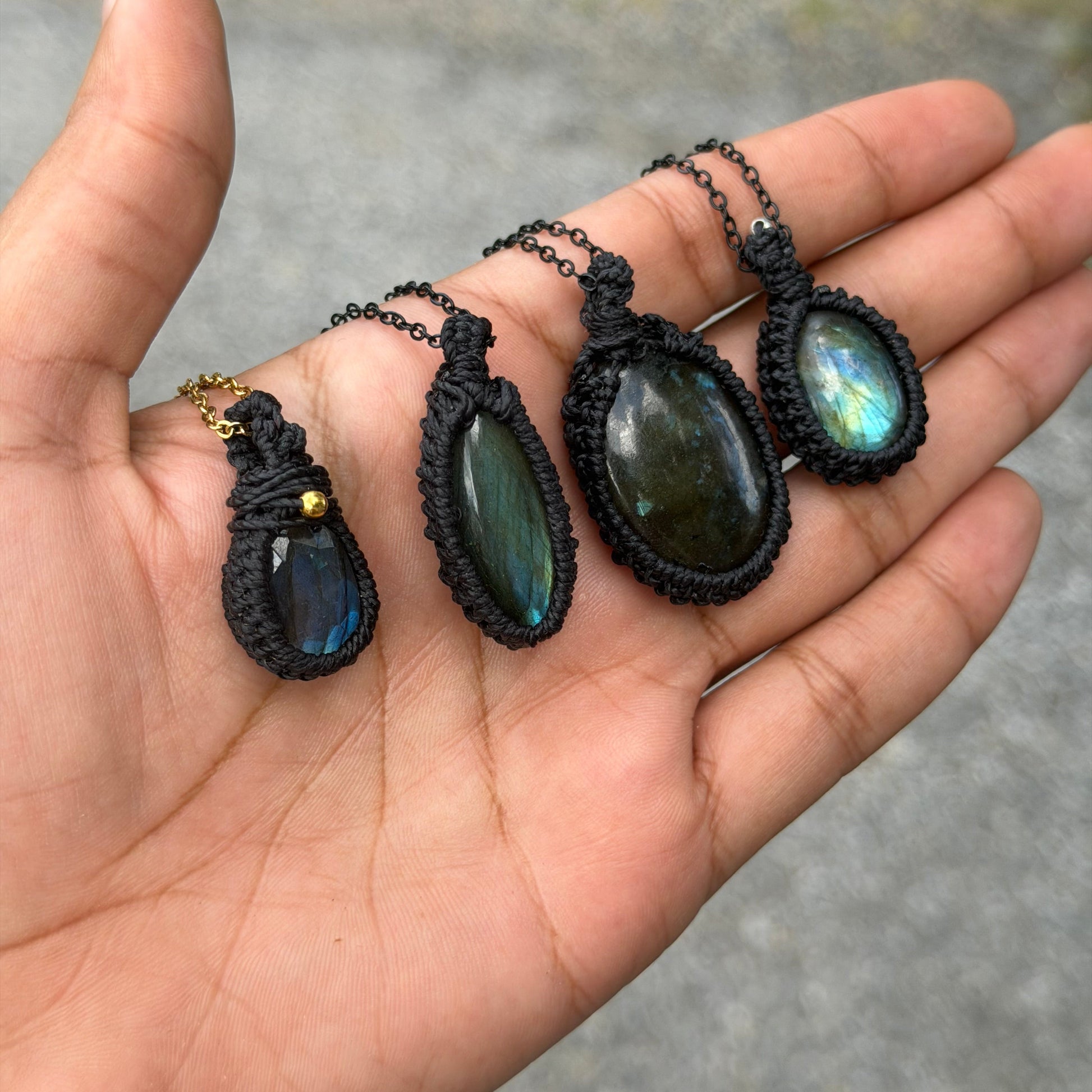 Three handmade labradorite pendants in black macrame wraps are displayed on an open hand outdoors.