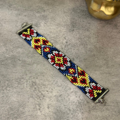 A beaded bracelet with a geometric diamond pattern in blue, red, yellow, and white, featuring silver-toned metal clasp ends, displayed on a textured gray surface.
