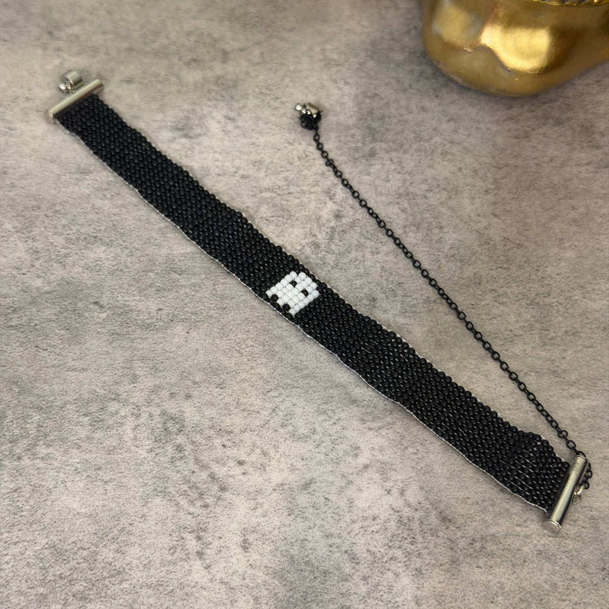 A black beaded bracelet with a small white geometric charm in the center, displayed on a textured gray surface next to a gold decorative object.