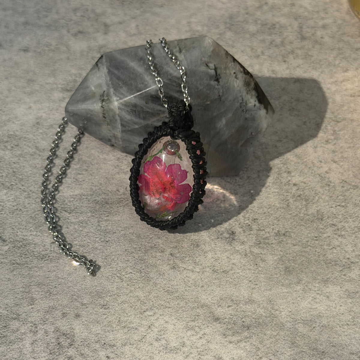 A handmade oval pendant necklace featuring a pink flower preserved inside clear resin, set in a black braided cord frame and hanging from a silver chain, displayed on a gray stone against a textured gray background.