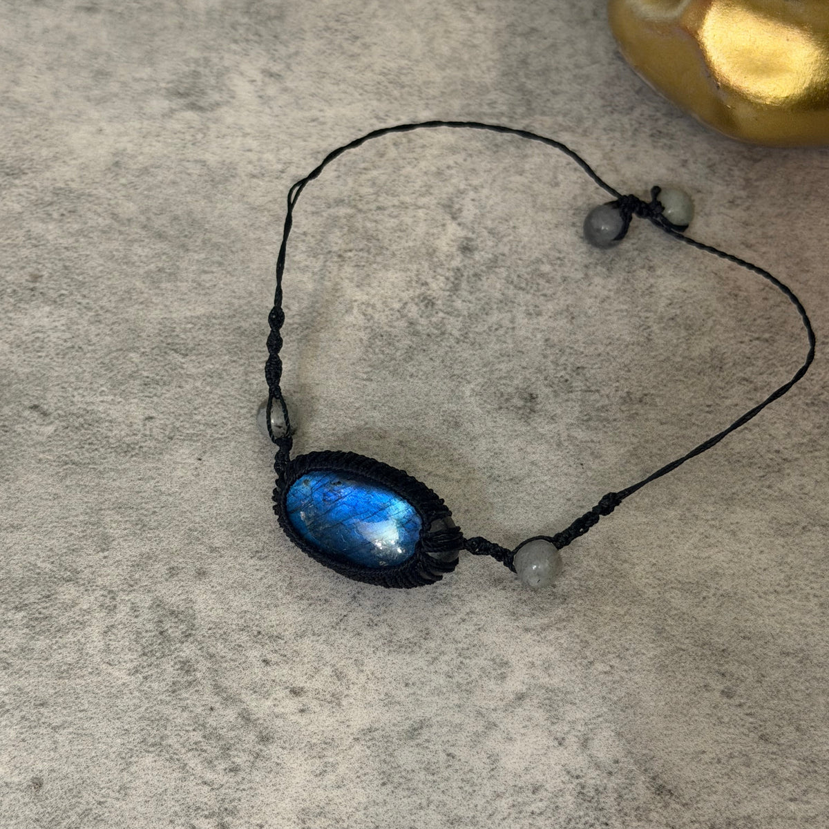 A black macrame bracelet featuring a central oval pendant with a shimmering blue and green iridescent stone, accented with small gray beads, displayed on a textured gray surface.