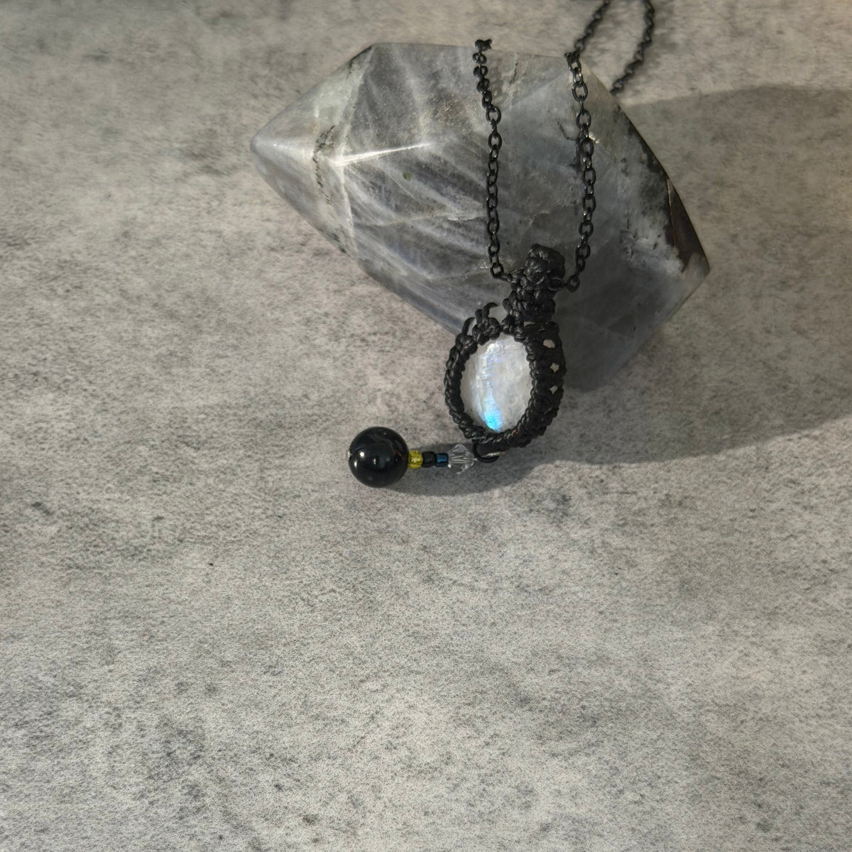 A black macrame pendant necklace with a white moonstone and a small black bead charm, displayed on a textured gray surface next to a clear crystal.