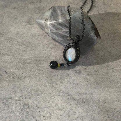 A black macrame pendant necklace with a white moonstone and a small black bead charm, displayed on a textured gray surface next to a clear crystal.