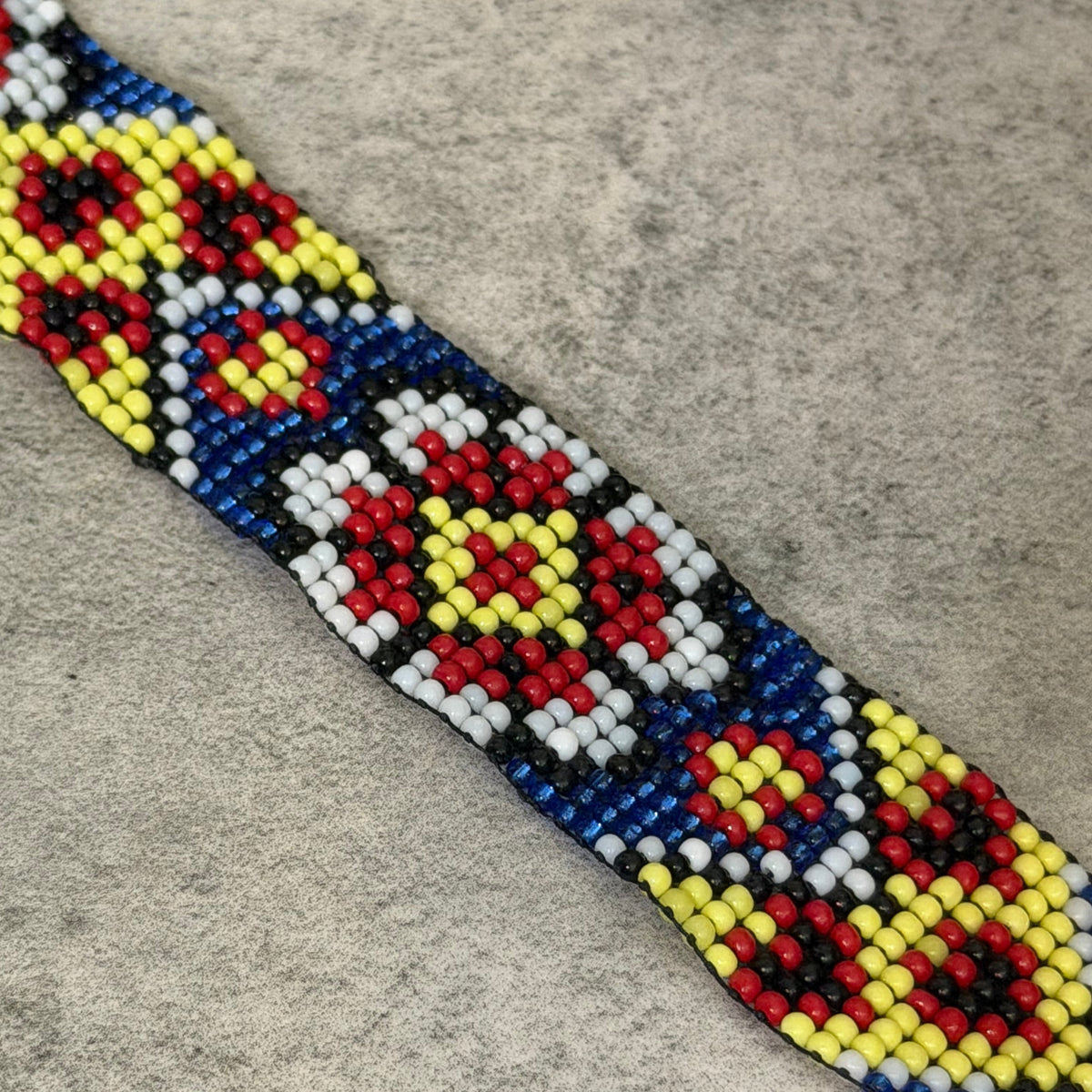 A close-up of a beaded belt or strap featuring a geometric pattern with red, yellow, black, white, and blue seed beads on a textured gray background.