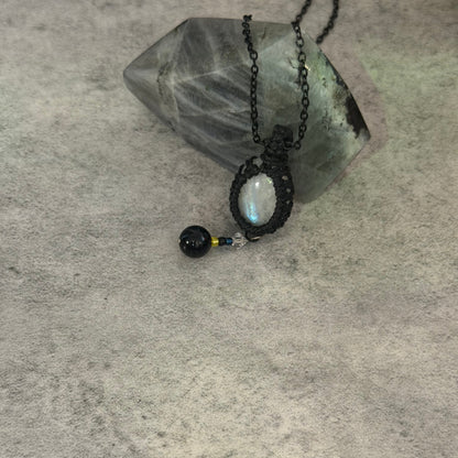 A black chain necklace with a handmade black macrame pendant holding an oval iridescent moonstone, displayed on a gray textured surface next to a crystal cluster.
