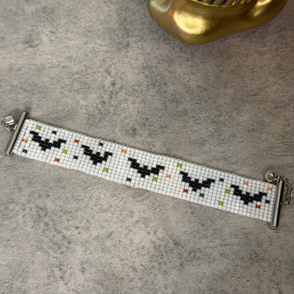 A beaded bracelet with a white mesh band, black bat motifs, and small multicolor dot accents, displayed on a gray textured surface next to a gold skull prop.