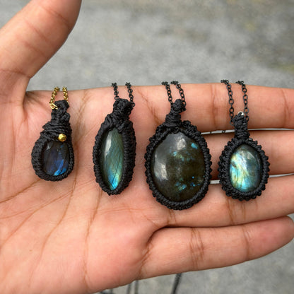 Four handmade labradorite pendant necklaces with black macrame wraps are displayed on an open hand, set against a blurred outdoor background.