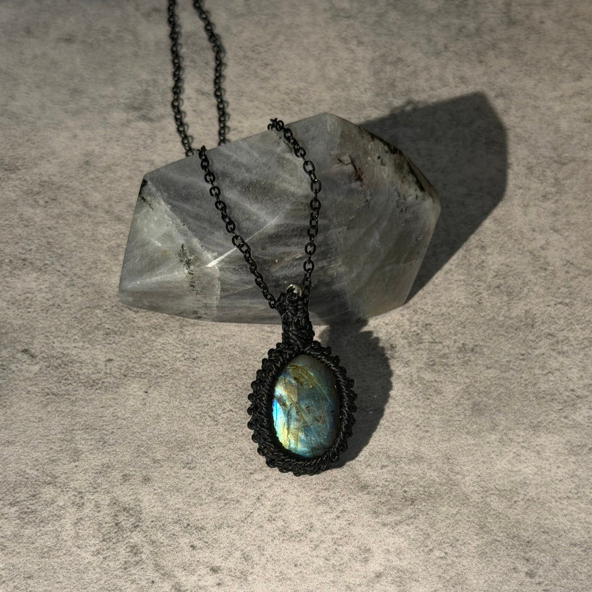 A black macrame pendant necklace with an oval iridescent labradorite stone, displayed on a textured gray surface with a clear quartz crystal.