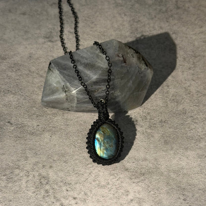 A black macrame pendant necklace with an oval iridescent labradorite stone, displayed on a textured gray surface with a clear quartz crystal.