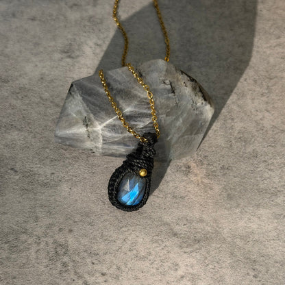 A teardrop pendant necklace with a black macrame-wrapped setting and a blue iridescent labradorite stone, hanging from a gold chain and resting on a clear quartz crystal against a textured gray background.
