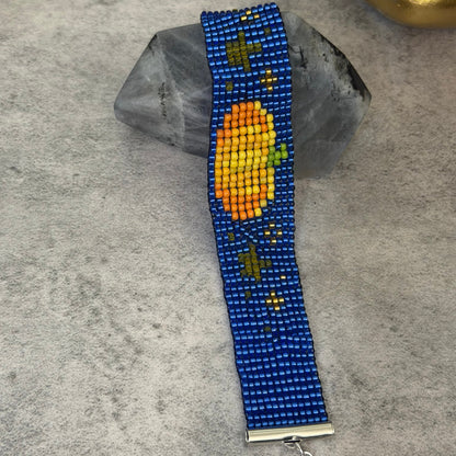 A beaded bracelet with a blue base, featuring a central orange and yellow pumpkin design with small green leaves and stars, displayed on a gray stone against a textured gray background.