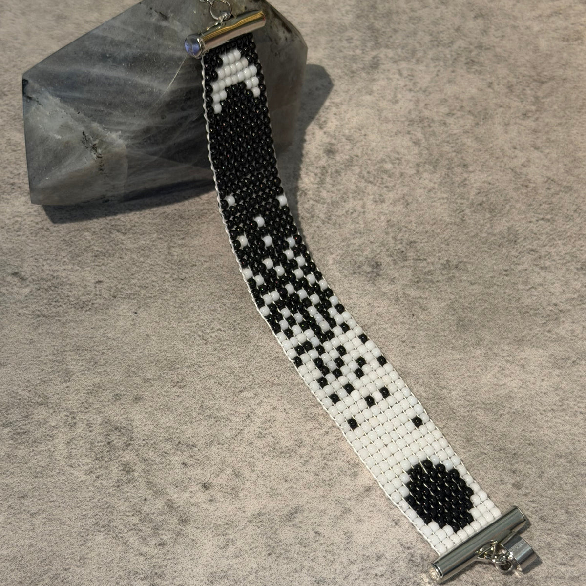 A handmade beaded watch band with a black and white geometric pattern, resting on a textured gray surface next to a polished crystal.