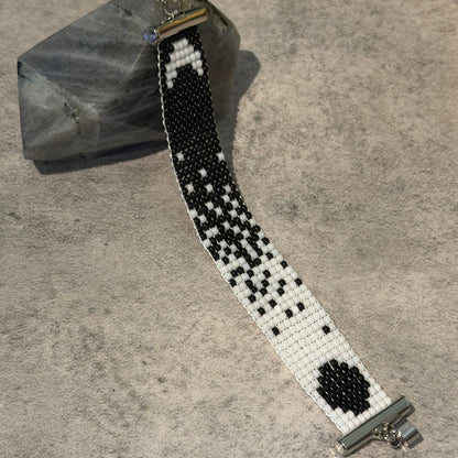 A handmade beaded watch band with a black and white geometric pattern, resting on a textured gray surface next to a polished crystal.