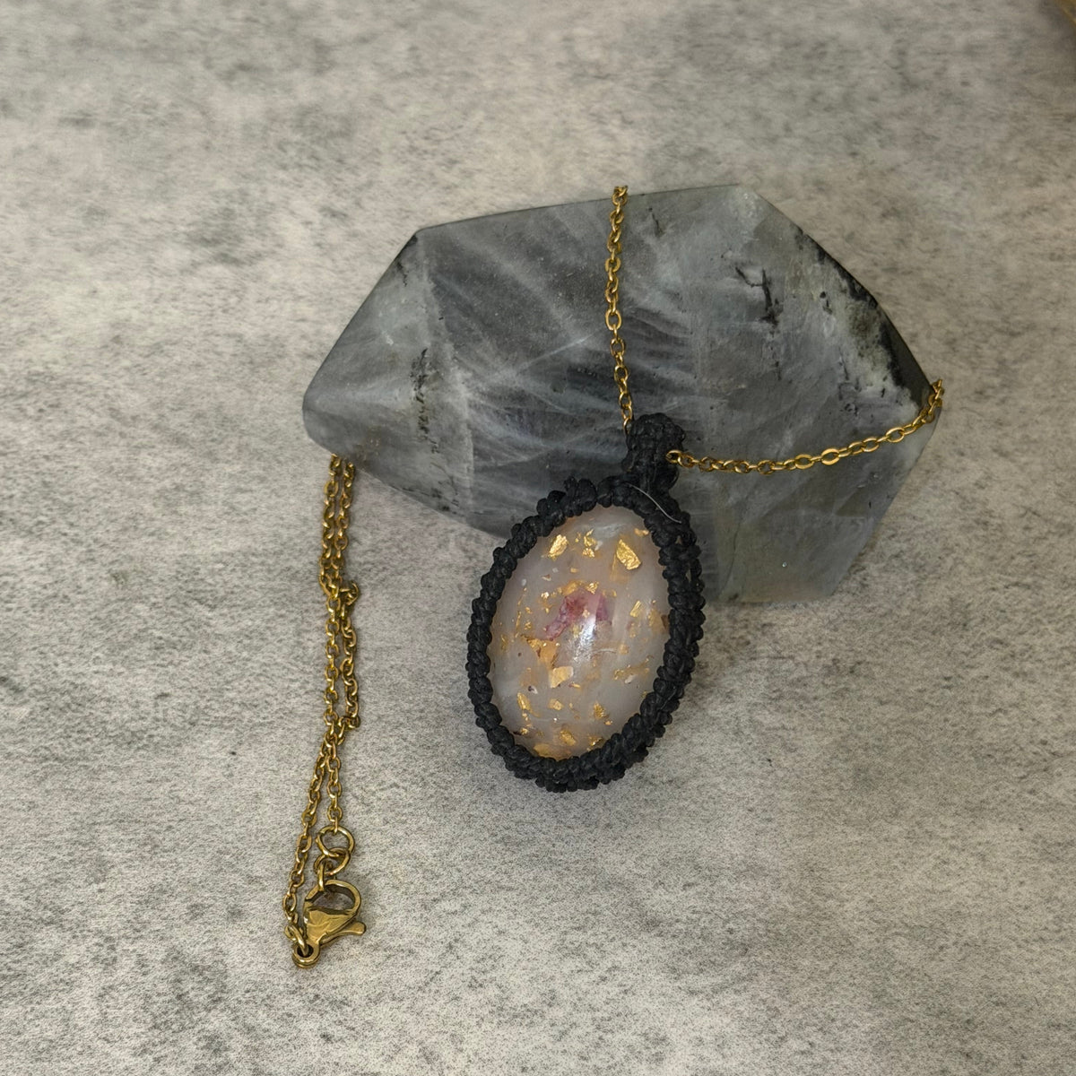 A handmade pendant necklace featuring an oval light gray stone with gold flecks, set in a black macrame border and suspended from a gold chain, displayed on a gray crystal cluster over a textured gray surface.