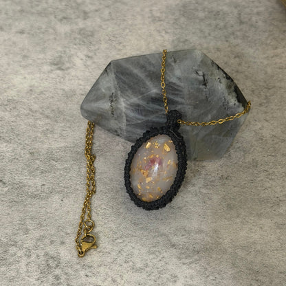 A handmade pendant necklace featuring an oval light gray stone with gold flecks, set in a black macrame border and suspended from a gold chain, displayed on a gray crystal cluster over a textured gray surface.