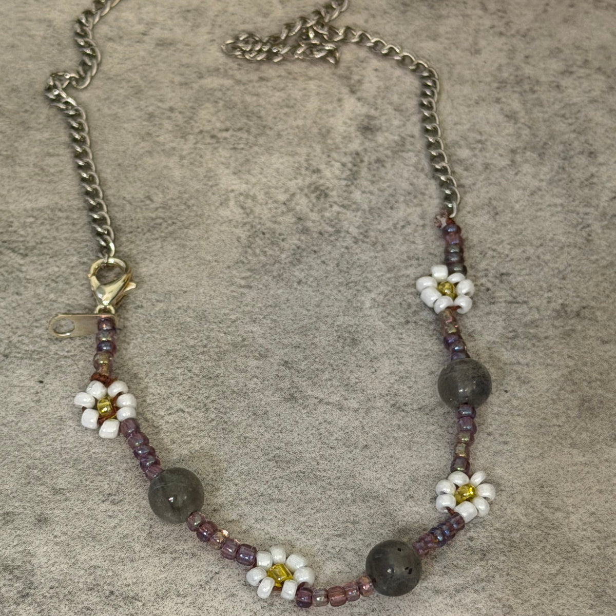A beaded necklace featuring white daisy flowers with yellow centers, dark gray round beads, and small purple beads, suspended from a silver-toned chain on a textured gray background.