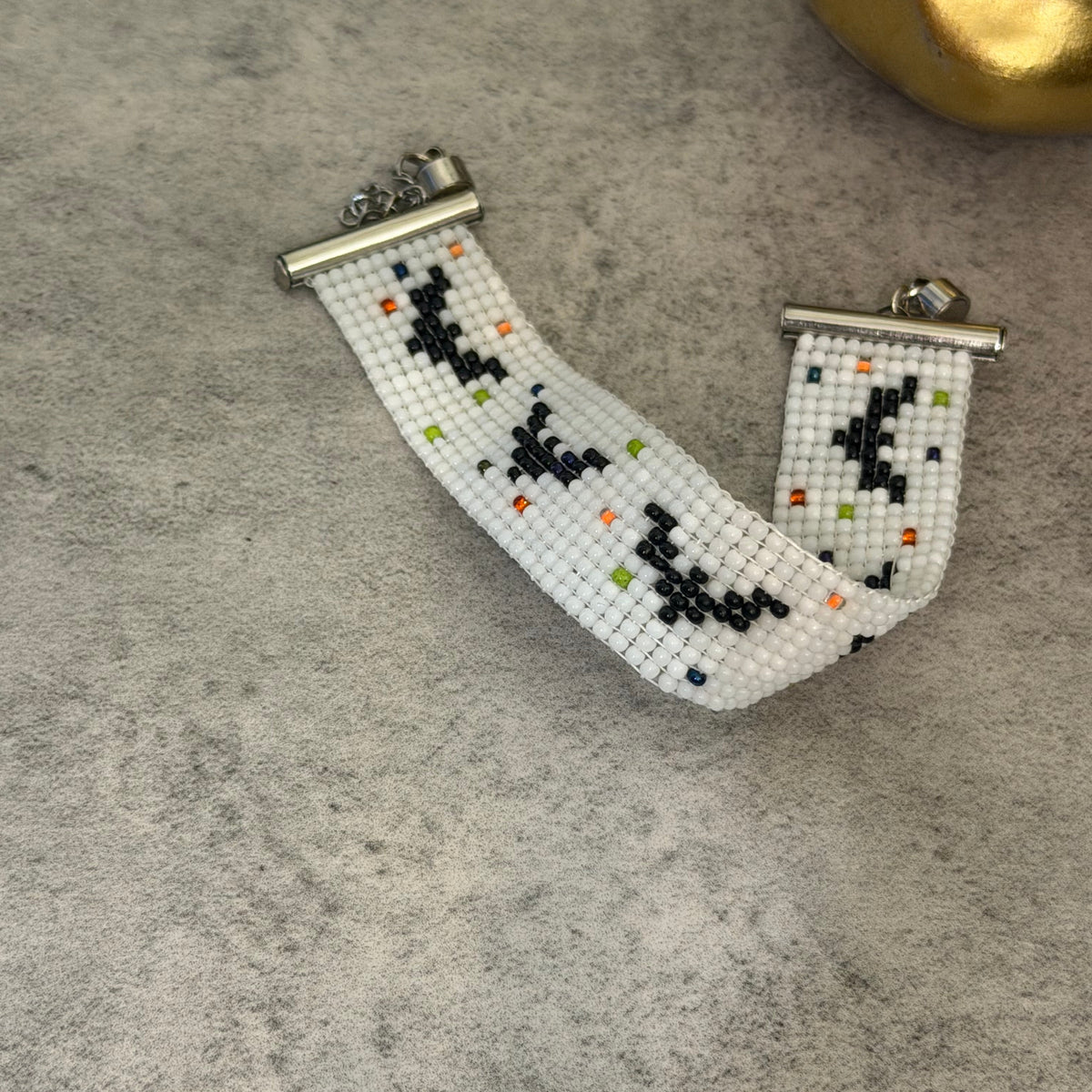 A handmade beaded bracelet with a white base, black geometric patterns, and small multicolor accent beads, resting on a textured gray surface.