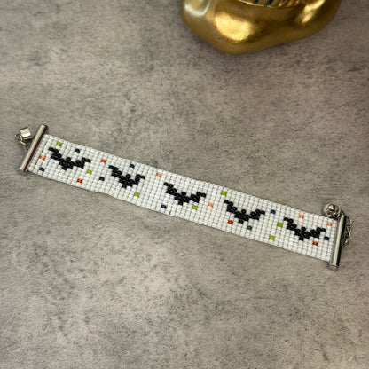 A beaded bracelet featuring a white base with black bat motifs and small multicolored dots, displayed on a gray textured surface with a gold skull prop in the background.