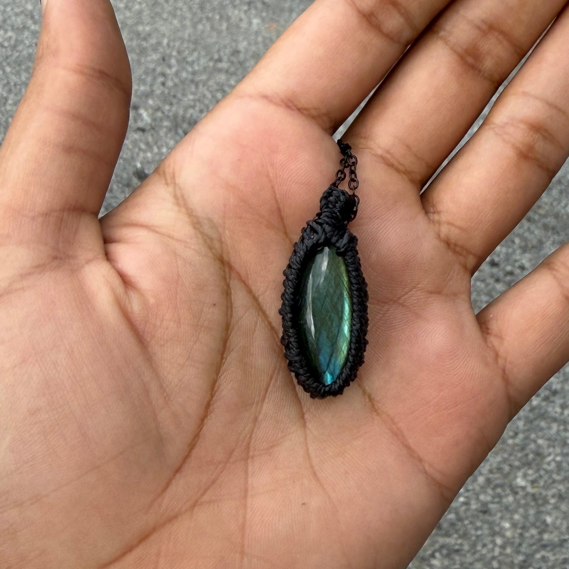 A hand holding a small black macrame pendant with an oval iridescent green-blue stone, photographed outdoors against a gray paved background.