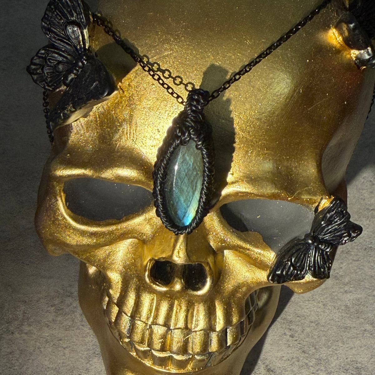 A gothic-style pendant necklace featuring a large oval iridescent blue-green stone set in black metal, displayed on a gold skull mannequin with black butterfly embellishments.