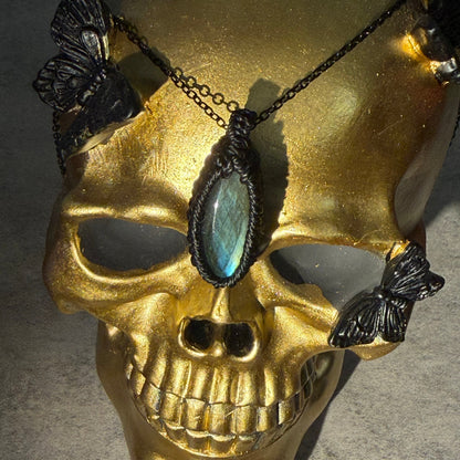 A gothic-style pendant necklace featuring a large oval iridescent blue-green stone set in black metal, displayed on a gold skull mannequin with black butterfly embellishments.