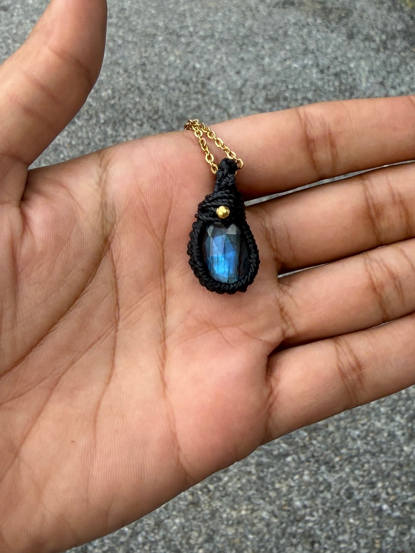 A close-up of a hand holding a teardrop-shaped pendant necklace. The pendant features a black macrame-style wrap around a blue iridescent stone, suspended from a gold chain.