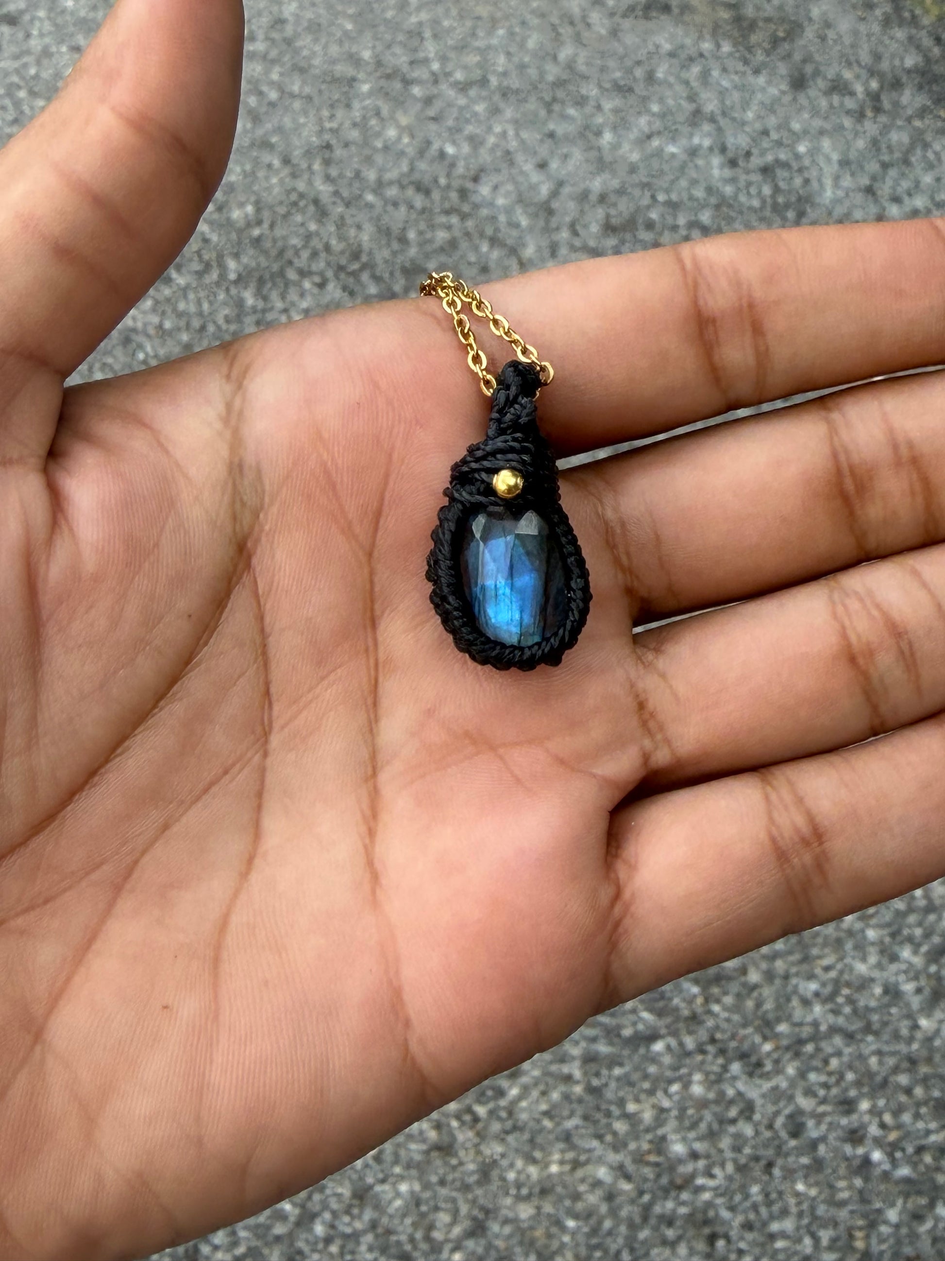 A close-up of a hand holding a teardrop-shaped pendant necklace. The pendant features a black macrame-style wrap around a blue iridescent stone, suspended from a gold chain.