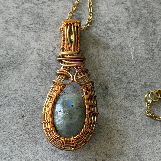 A handmade pendant necklace featuring a round labradorite stone wrapped in textured copper wire, resting on a gray textured surface.