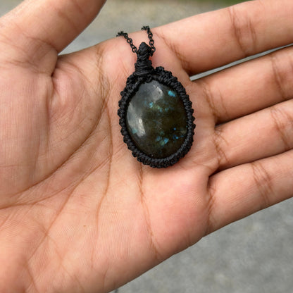A handmade oval pendant with a dark green and blue marbled stone, wrapped in black macrame, held in a person's hand against an outdoor blurred background.