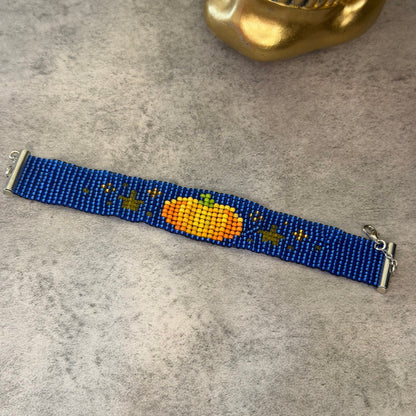 A beaded stretch bracelet featuring a blue base with a central orange pumpkin design and small gold stars, displayed on a gray textured surface with a gold skull prop in the background.