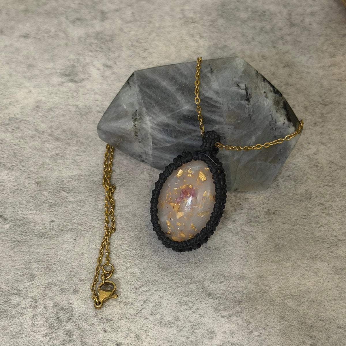 A handmade pendant necklace featuring a black macrame-wrapped oval bezel with a white opal containing gold flakes, resting on a gray crystal cluster against a textured gray background.