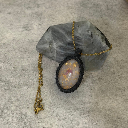 A handmade pendant necklace featuring a black macrame-wrapped oval bezel with a white opal containing gold flakes, resting on a gray crystal cluster against a textured gray background.