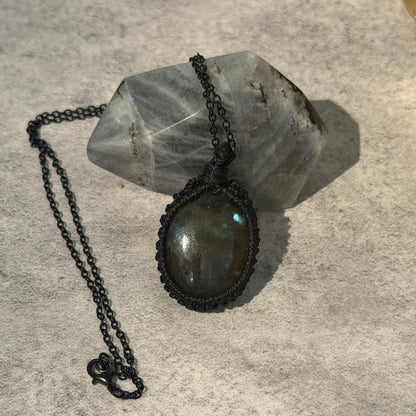 A handmade oval pendant necklace featuring a dark gray labradorite stone in a black woven cord and metal setting, resting on a gray crystal cluster against a textured gray background.