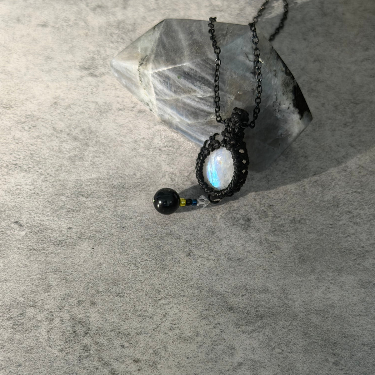 A handmade black macrame pendant necklace with a round iridescent moonstone and a small black bead charm, displayed on a textured gray surface next to a clear crystal.