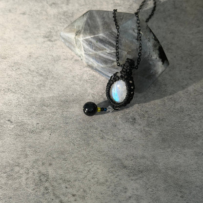 A handmade black macrame pendant necklace with a round iridescent moonstone and a small black bead charm, displayed on a textured gray surface next to a clear crystal.