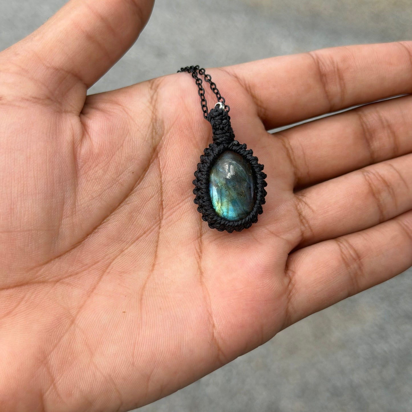 A hand holding a black macrame pendant necklace featuring an oval labradorite stone with a blue-green iridescent sheen, set against an outdoor blurred background.