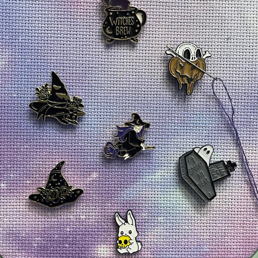 A collection of gothic-themed magnetic enamel needle minders displayed on a fabric surface, with designs including a witch's hat and other related motifs.